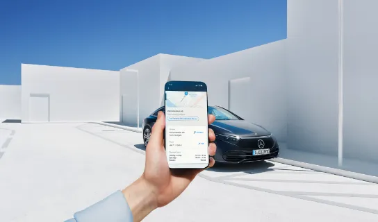 Eine Hand hält ein Smartphone mit der Mercedes-Benz App. Im Hintergrund ist ein schwarzes Mercedes-Benz Fahrzeug vor weißen Wänden zu sehen.