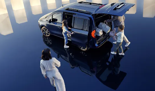 Der Kofferraum eines dunkelblauen Mercedes-Benz EQT wird von einem Mann und einem Jungen beladen. Ein Mädchen öffnet die Schiebetür und eine Frau geht auf das Fahrzeug zu.