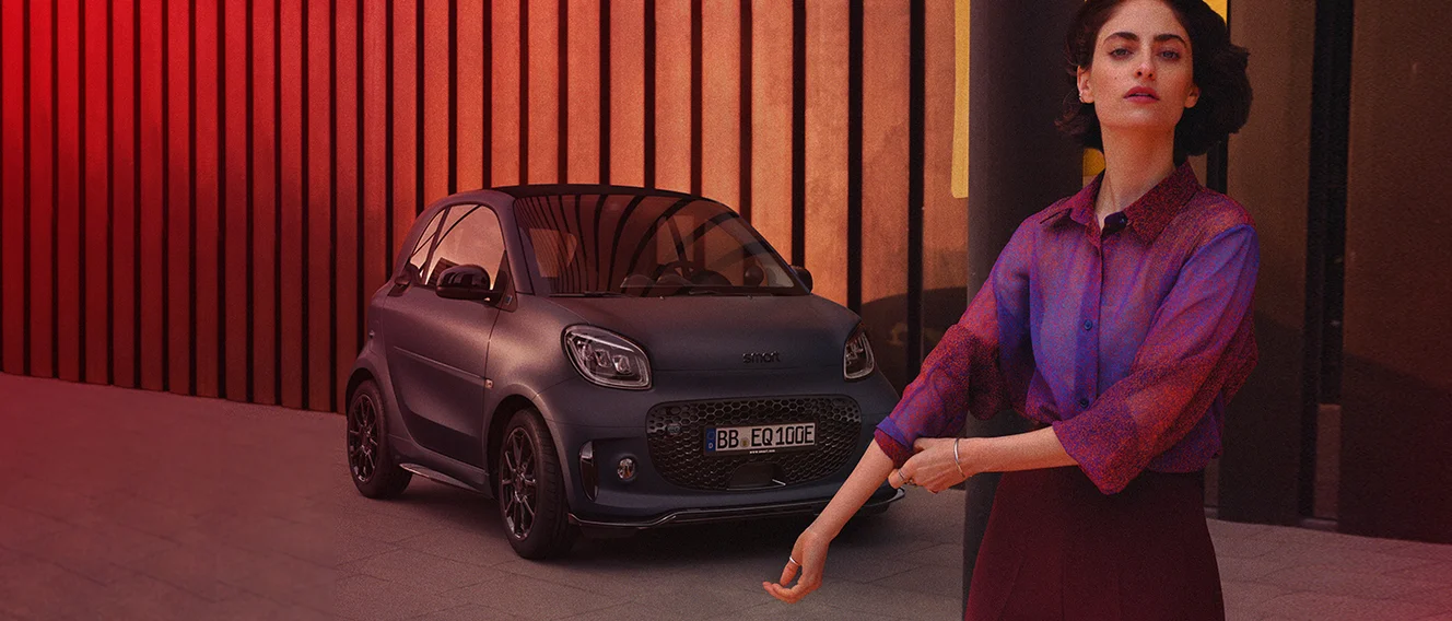 Ein dunkelgrauer smart fortwo steht rot beleuchtet vor einer Lamellenwand, vor ihm steht eine Frau mit lila Bluse.