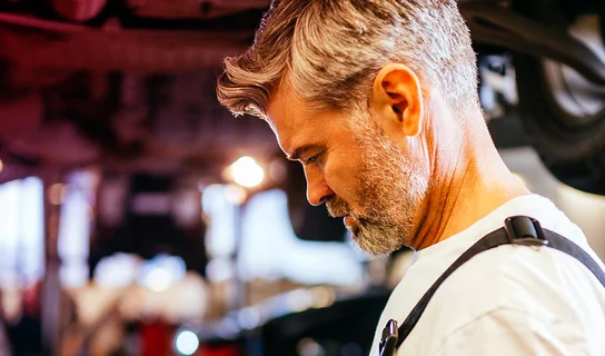 Ein Mechaniker arbeitet in einer Autowerkstatt. Im Hintergrund erkennt man ein Fahrzeug auf einer Hebebühne.