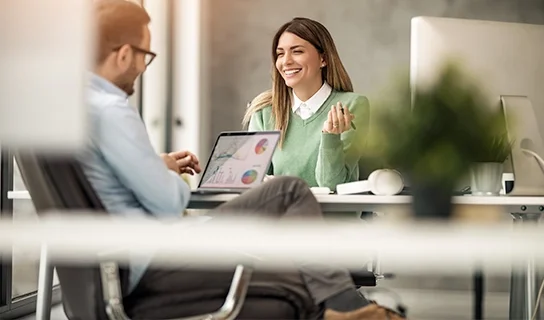 Ein Mann mit blauem Hemd und dunkelgrauer Hose sitzt am Laptop und eine Frau mit grünem Pullover und weißem Hemd sitzt ihm gegenüber am Tisch.